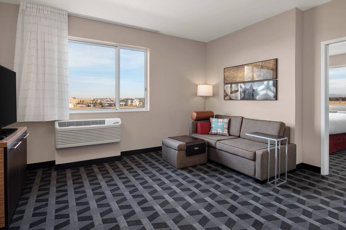 Een hotelkamer met een bank, bijzettafeltje, lamp, flatscreen televisie en een raam met uitzicht op de stad.