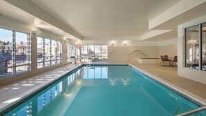 Uma grande piscina interior com paredes de vidro transparente, chão revestido em azulejos e área de estar com mesas e cadeiras.