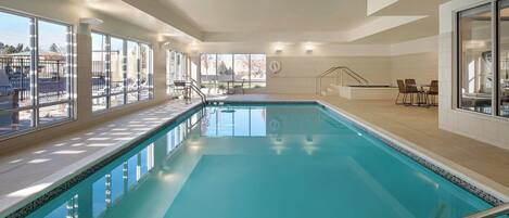 A large indoor swimming pool with clear glass walls, a tiled floor, and a seating area with tables and chairs.