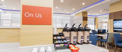 A coffee station with a sign that reads "On Us," a coffee dispenser, cups, and a bowl of oranges.