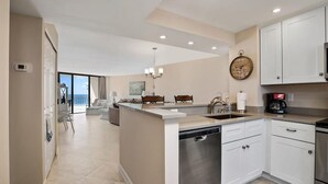 Cozinha moderna com armários brancos, lava-louças e cafeteira. Há uma área de jantar com mesa e cadeiras, e vista para o oceano através de amplas janelas.