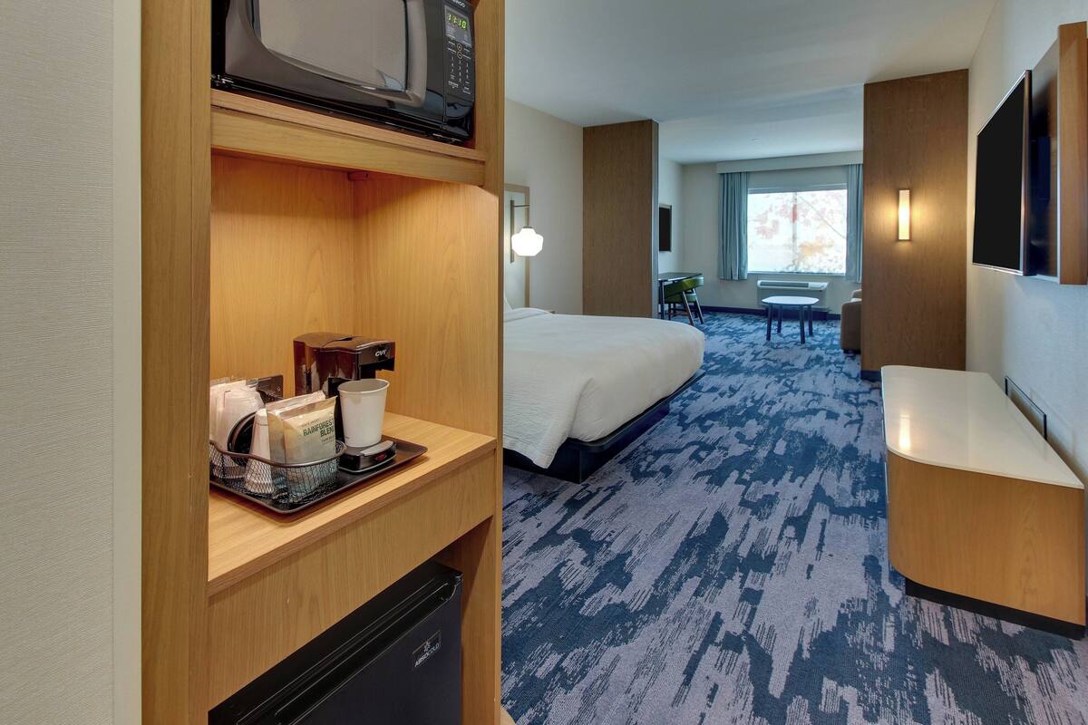 A hotel room with a bed, a desk with a coffee maker, a microwave, a TV, and a seating area with a chair and table.