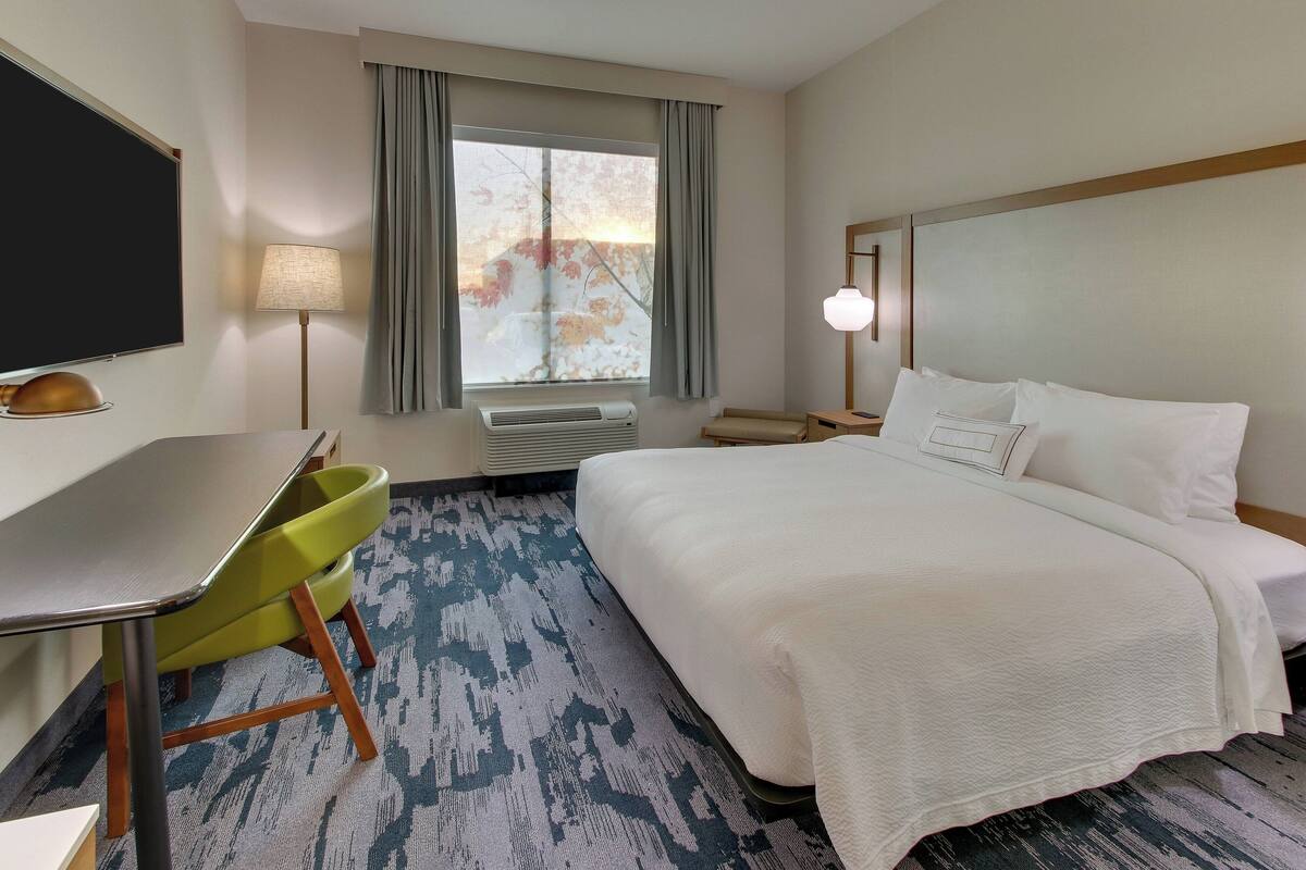 A hotel room with a bed, a desk, a chair, a TV, a window with curtains, and a lamp.