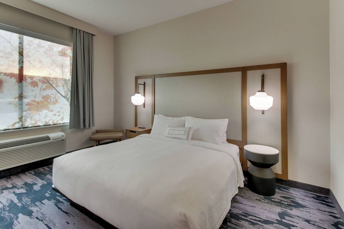 A hotel room with a large bed, two bedside lamps, a window with curtains, a bedside table, and a wall-mounted mirror.