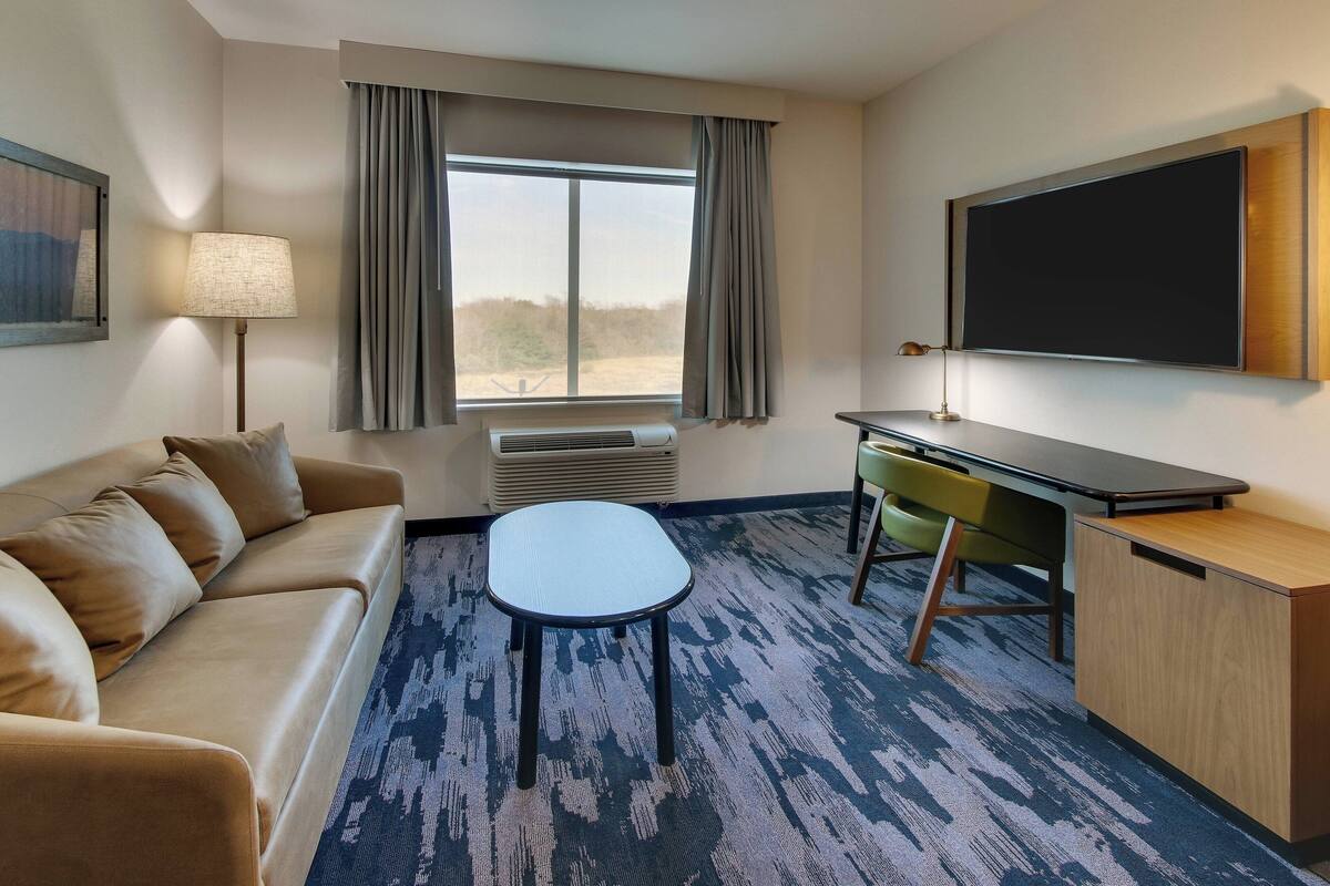 A hotel room with a sofa, a small table, a desk, a chair, a TV, and a window with curtains.