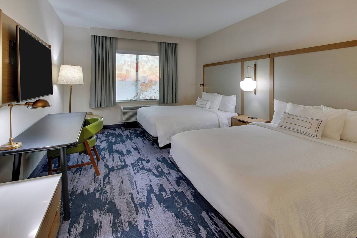 A hotel room with two beds, a desk, a chair, a TV, and a window with curtains.