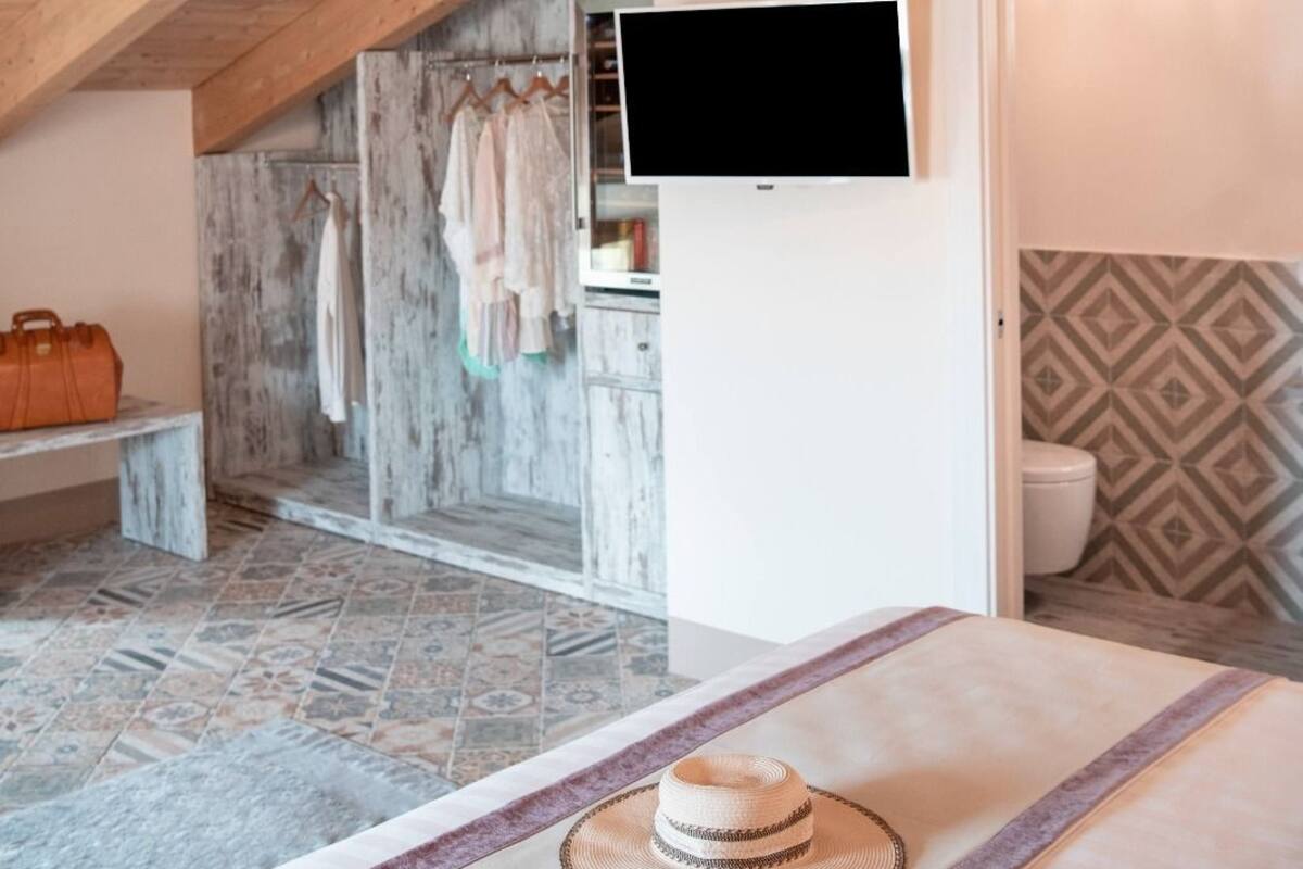 A bedroom with a wooden ceiling, a television mounted on the wall, a bed with a hat and a small bag, and a bathroom with patterned tiles.