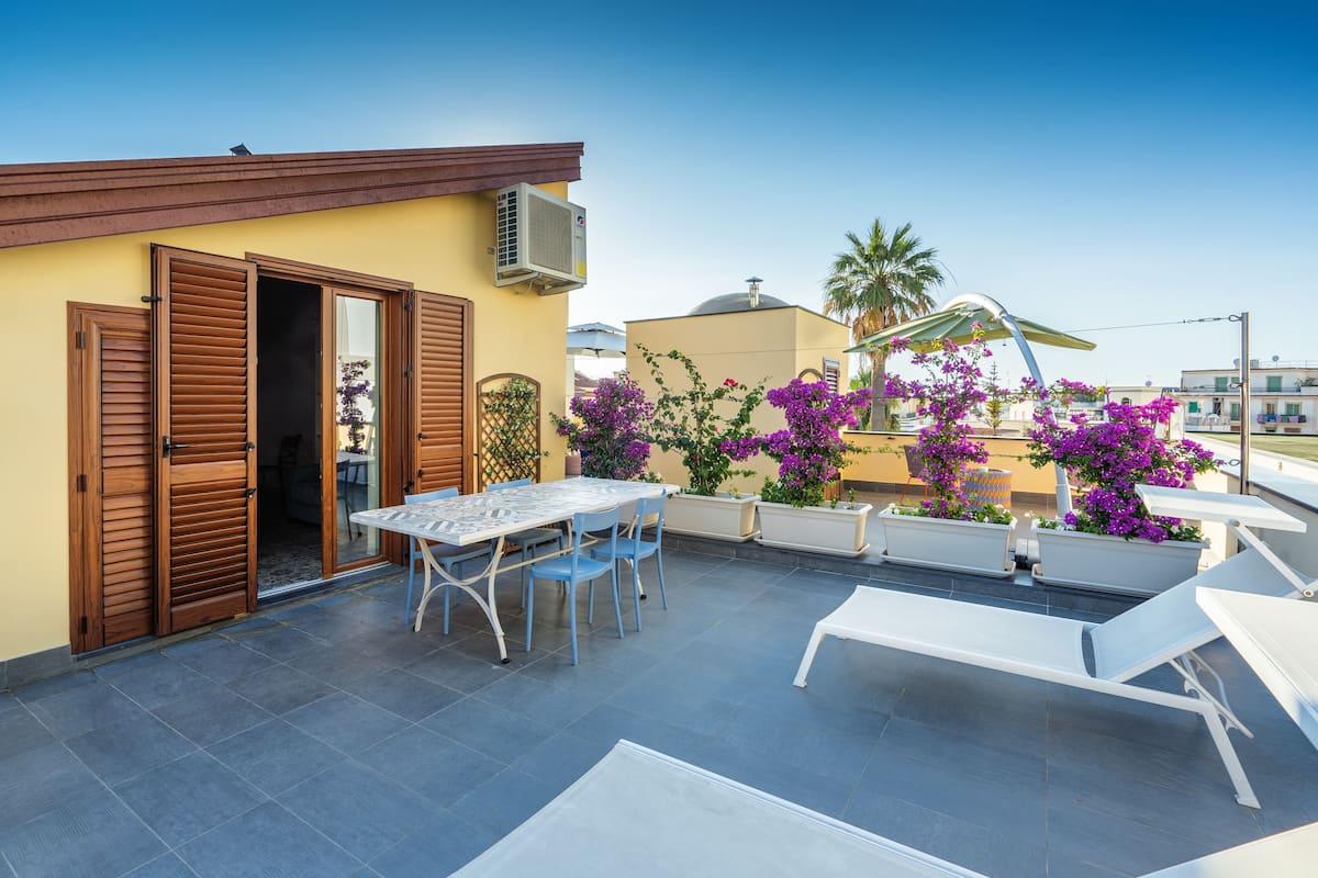 A rooftop terrace with a table and chairs, a white lounge chair, and potted plants with purple flowers.