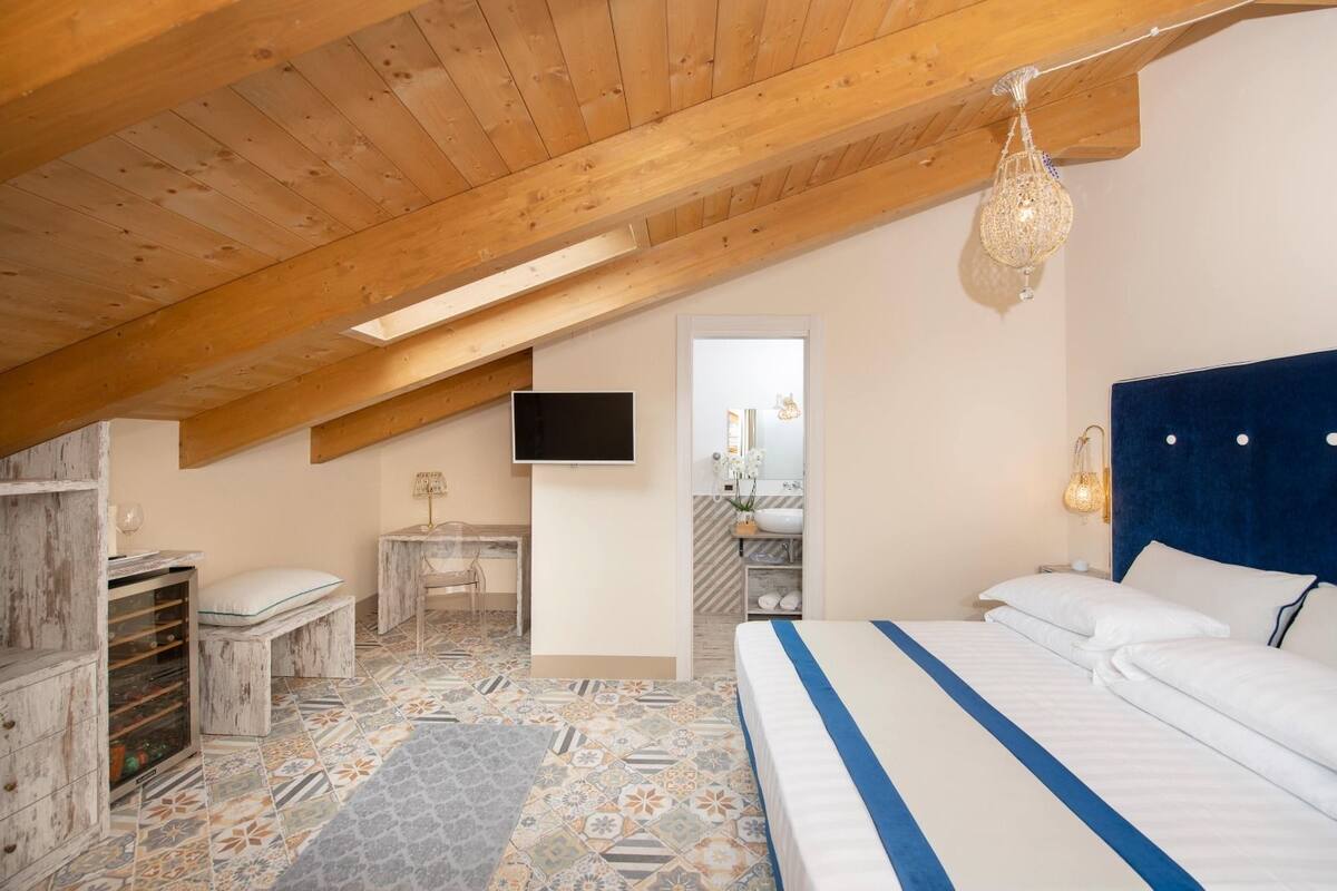 A bedroom with a sloped ceiling, a bed with a blue-striped bedspread, a desk with a chair, a television, and a bathroom visible through an open door.