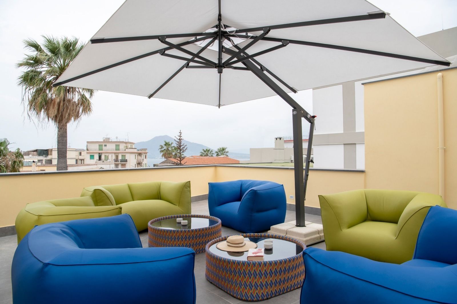 A rooftop terrace with blue and green sofas, a patterned ottoman, and a large umbrella.