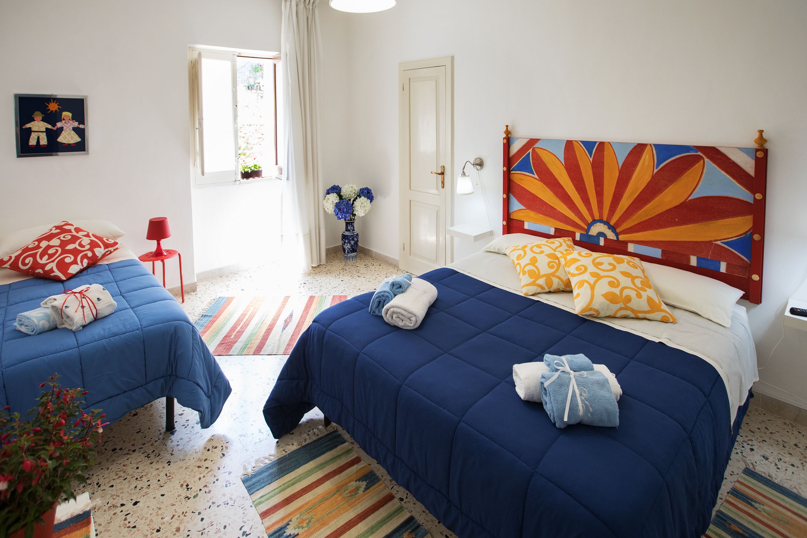 A hotel room with two beds, a colorful headboard, a window with curtains, a vase with flowers, and a striped rug.