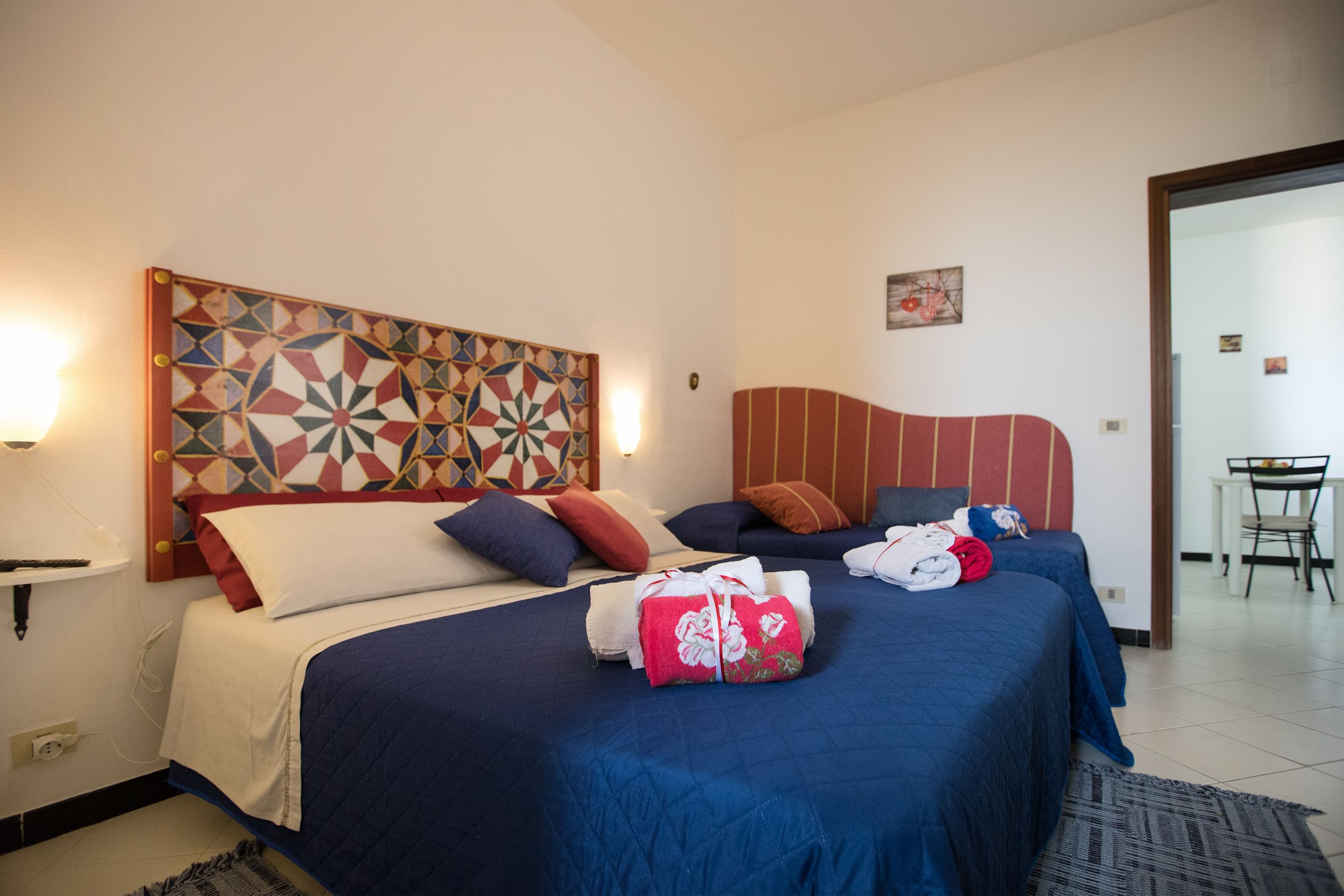 Una habitación de hotel con un cabecero grande de estampado geométrico, dos camas con ropa de cama azul y roja, y una pequeña mesa con sillas en la esquina.