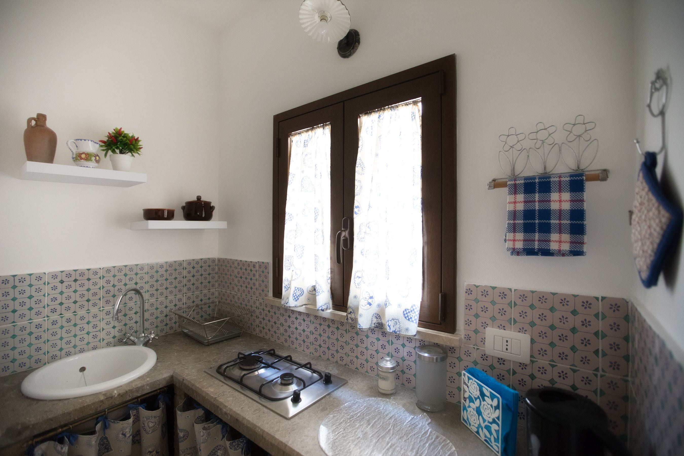 A kitchen with a sink, stove, and window with curtains. There are shelves with decorative items and a towel rack with a towel.