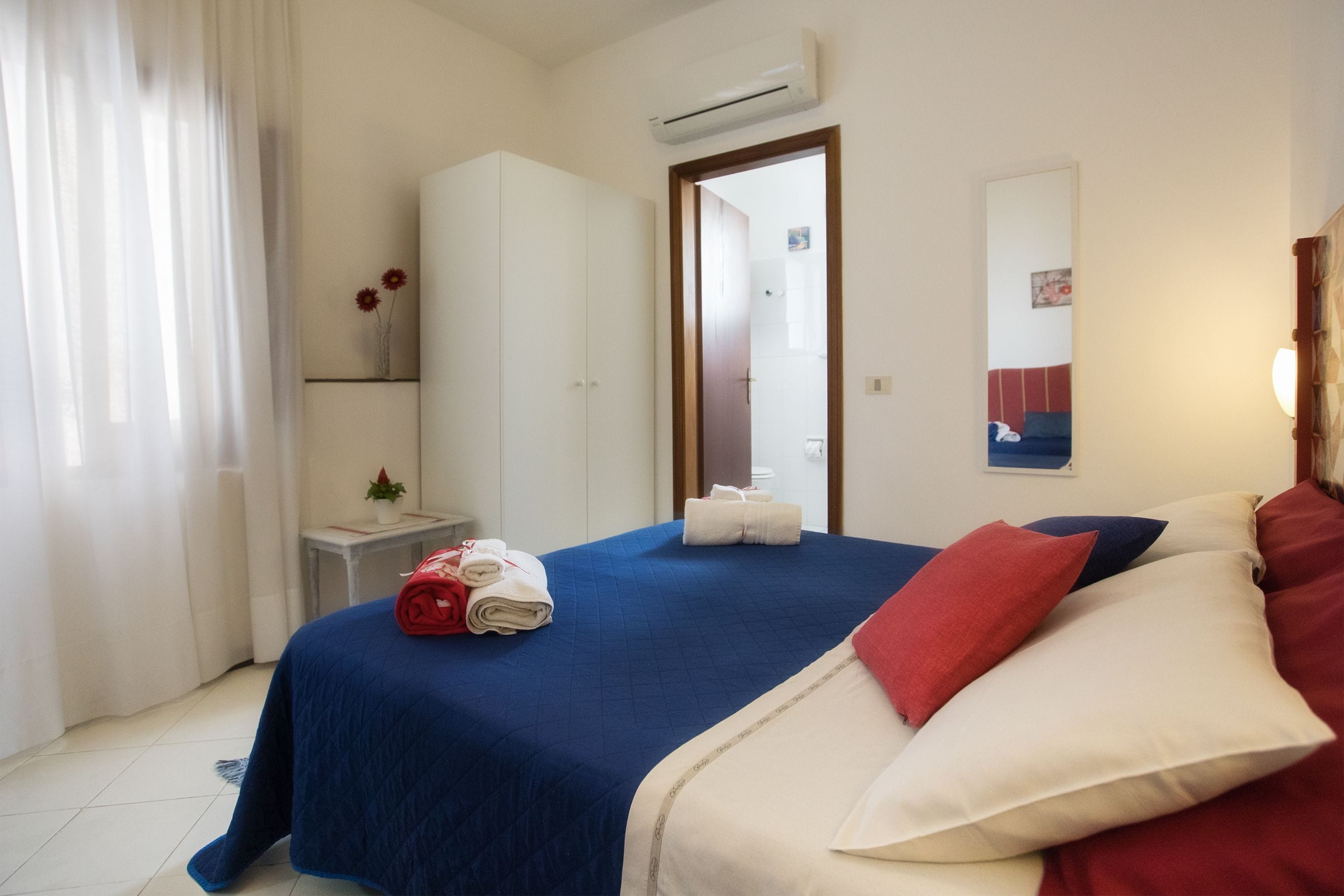 A hotel room with a bed, a wardrobe, a small table with a vase, and an air conditioning unit.