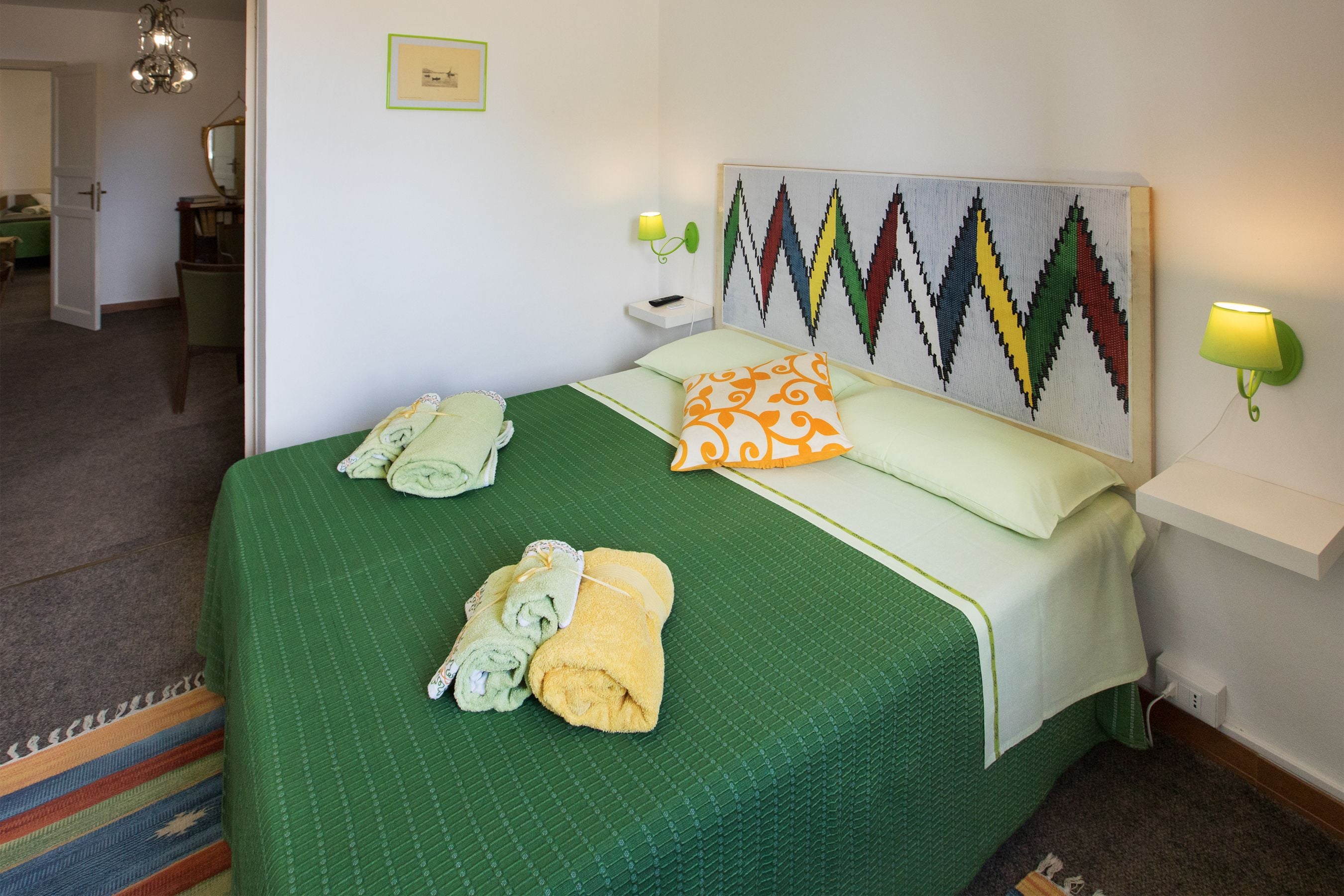 A neatly made bed with green bedding and two pillows, a wall-mounted artwork, and a bedside table with a lamp.