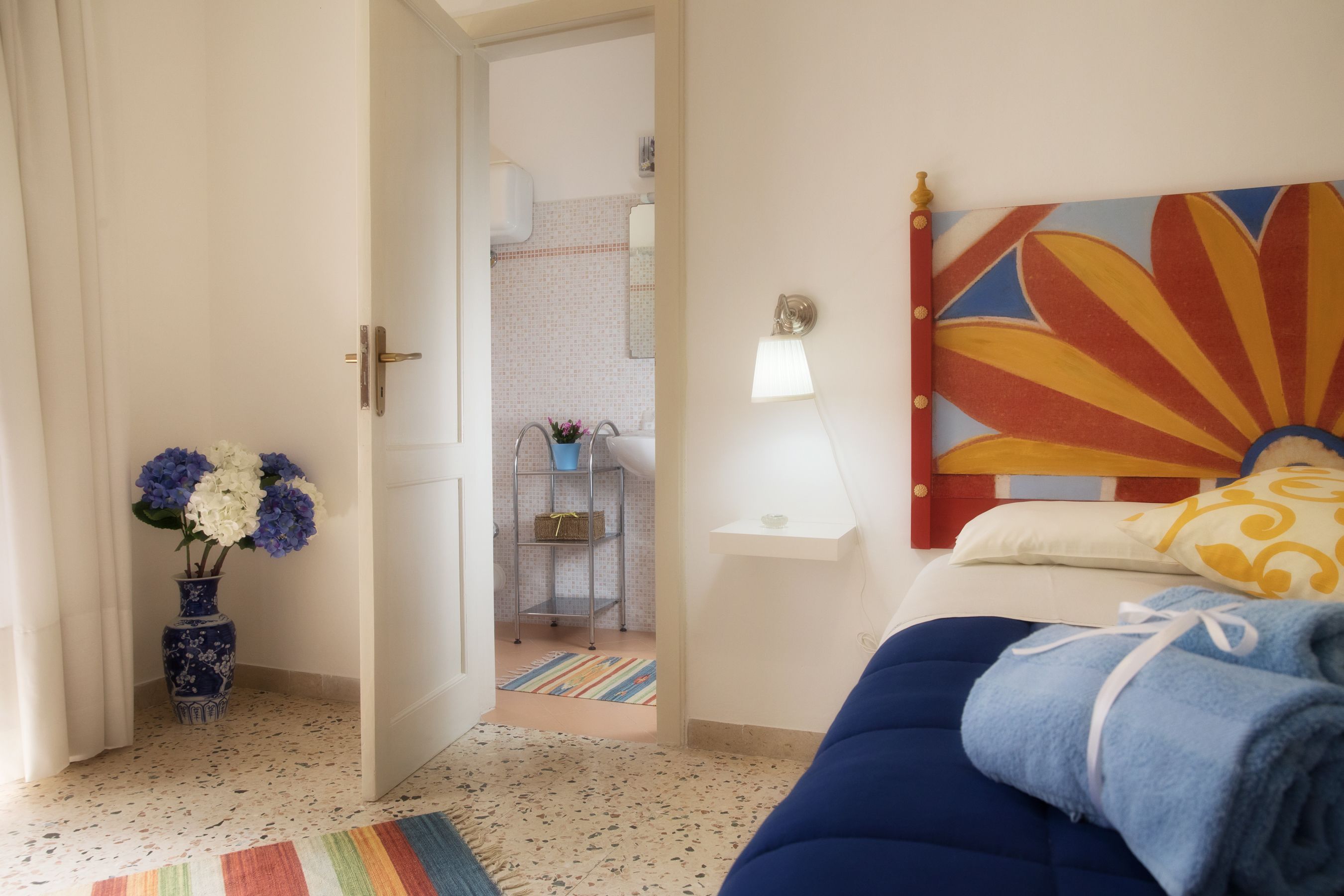 Ein Schlafzimmer mit Bett, einem farbenfrohen Bild an der Wand, einer Blumenvase und einem durchgegangenen Tür erkennbaren Badezimmer.