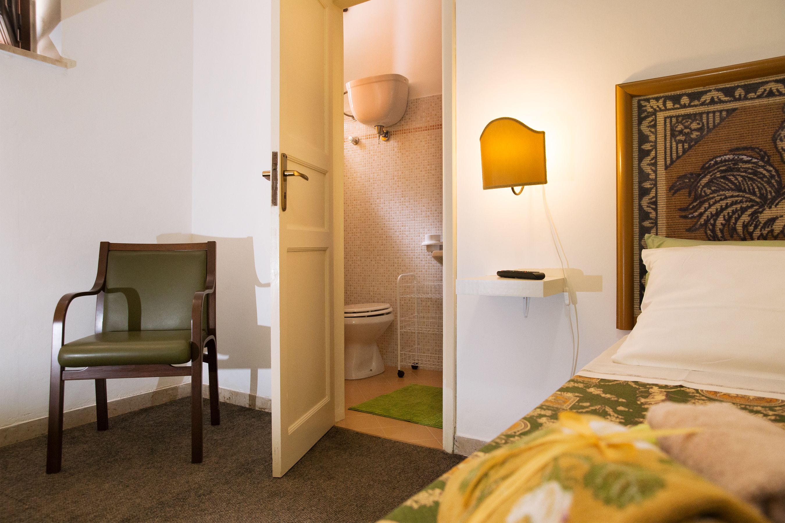 A hotel room with a bed, a chair, a bathroom with a toilet, and a wall-mounted lamp.