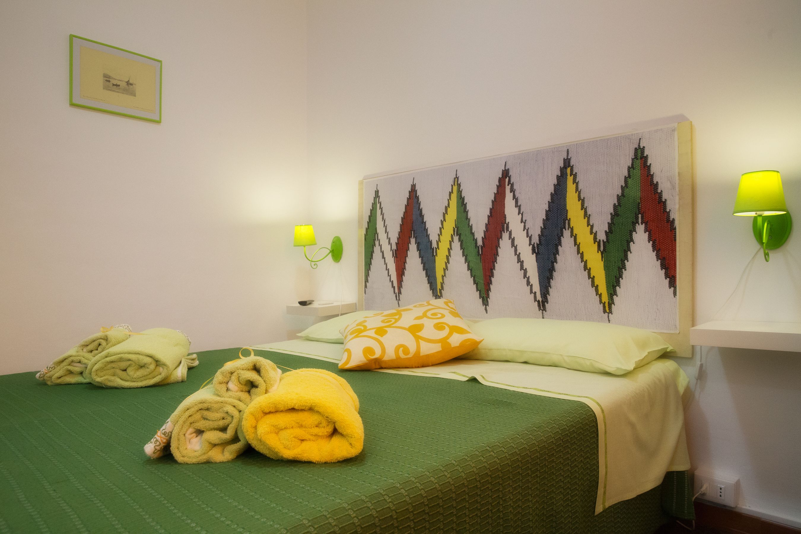 A neatly made bed with green bedding, a colorful wall tapestry, and two wall-mounted lamps.