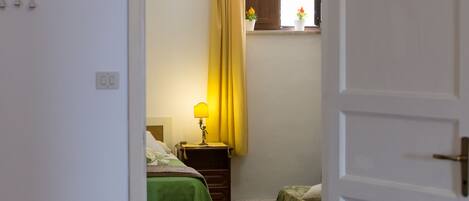 Un dormitorio con cama, una mesita de noche con lámpara, una ventana con cortinas y una pequeña mesa con una planta.