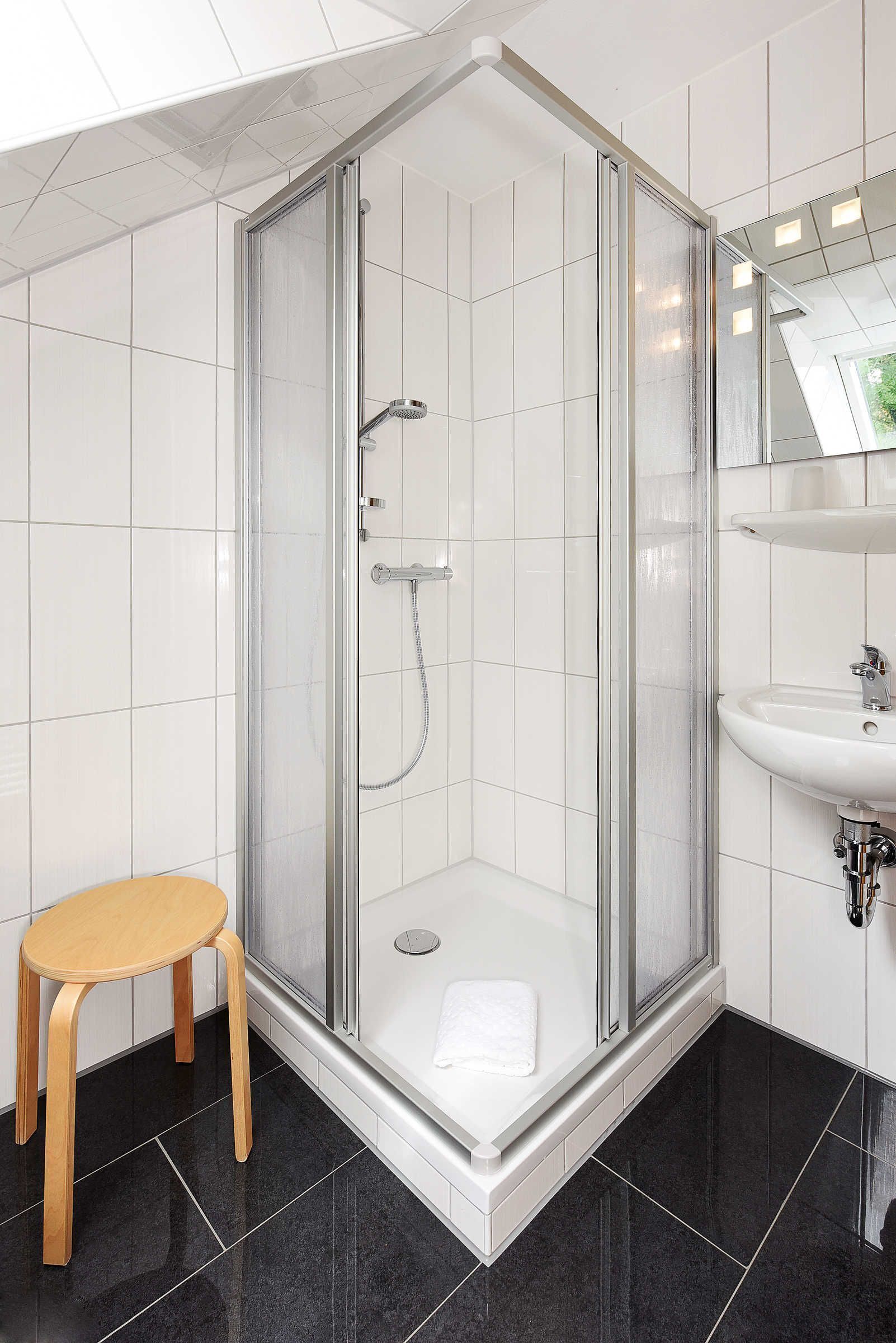 Eine Eckdusche mit Glastür, ein Holzschemel und ein weißes Waschbecken mit Spiegel darüber.