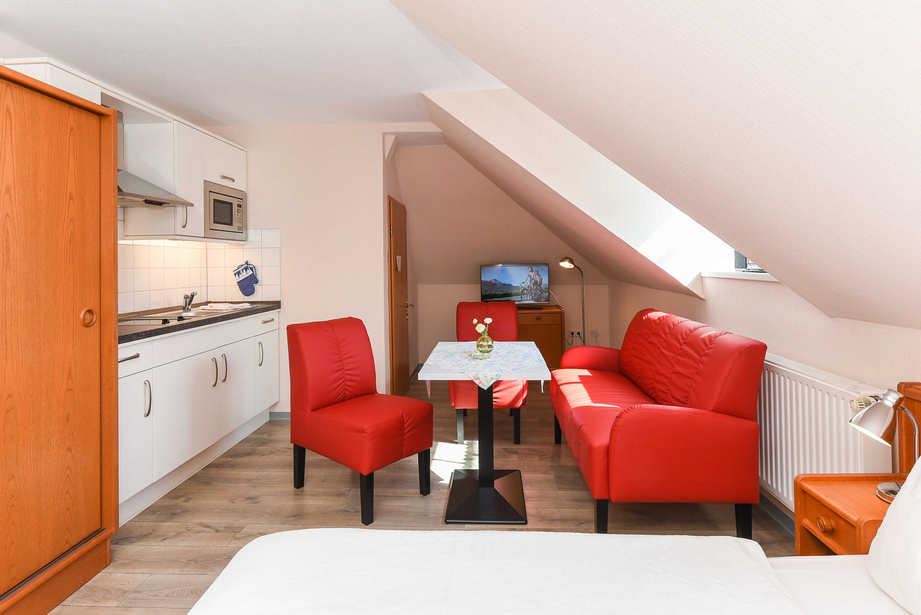 A compact room with a kitchenette, a dining area with red chairs, a small table, and a bed.