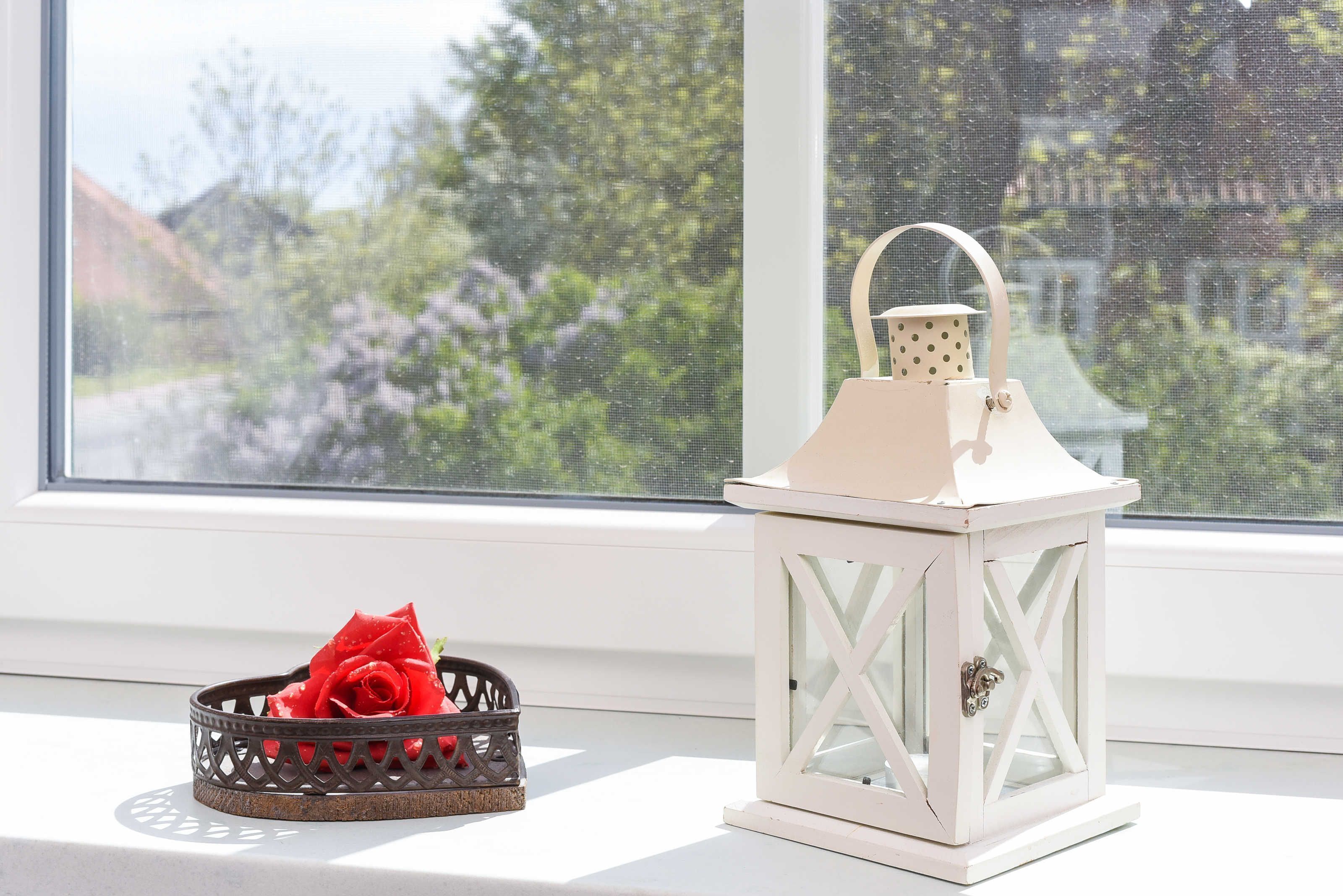 A white lantern with a handle on a windowsill next to a basket with a red rose.