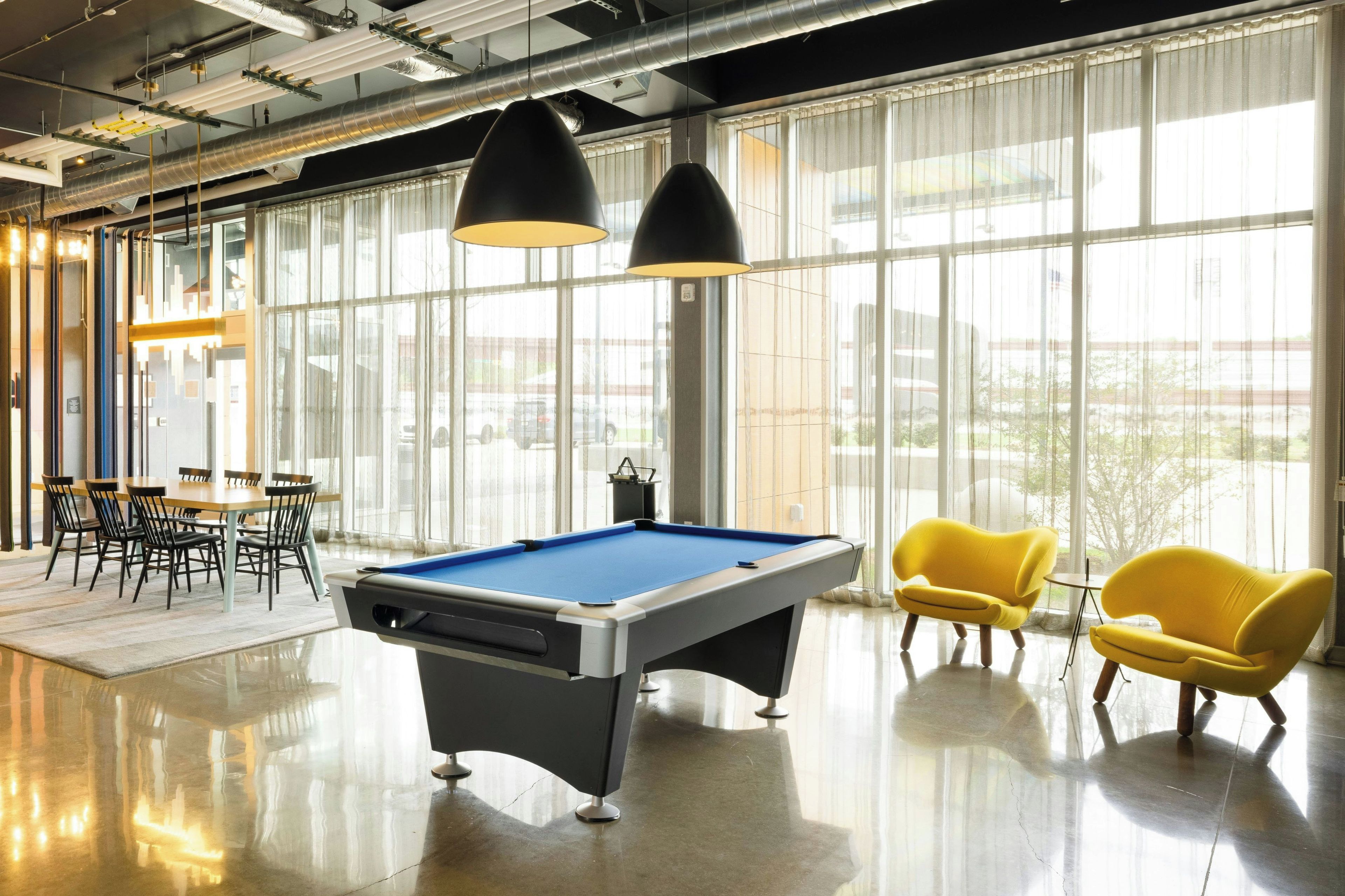 A billiards table with a pool cue, yellow chairs, and a table tennis table in a bright room with large windows.