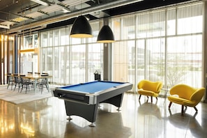 Una mesa de billar con taco, sillas amarillas y una mesa de tenis en una habitación luminosa con ventanas amplias.