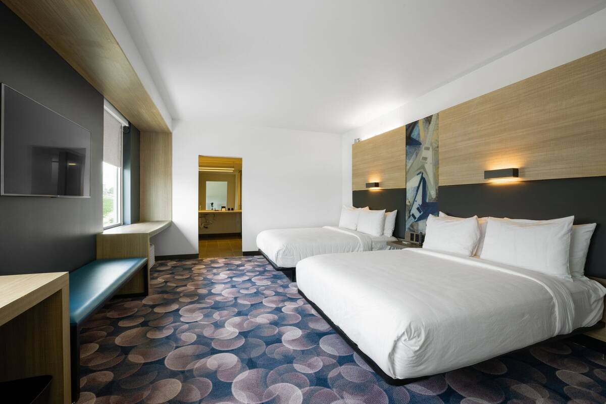 Habitación de hotel con dos camas, un escritorio, un banco, un ventanal grande y un cuadro en la pared.