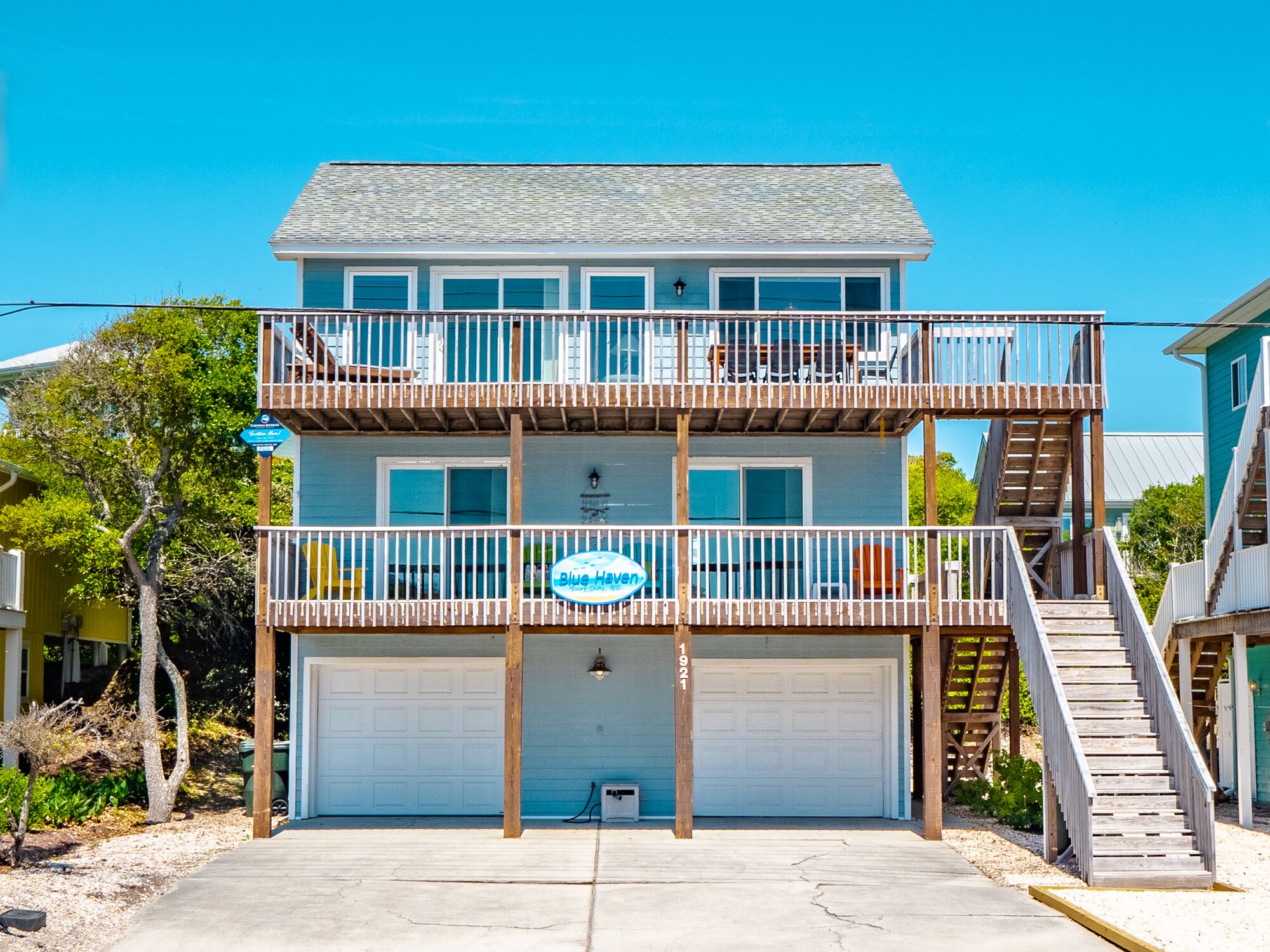 Ein zweistöckiges Strandhaus mit blauer Fassade, einer Holzterrasse und einem blauen Schild mit der Aufschrift 'Blue Haven'.