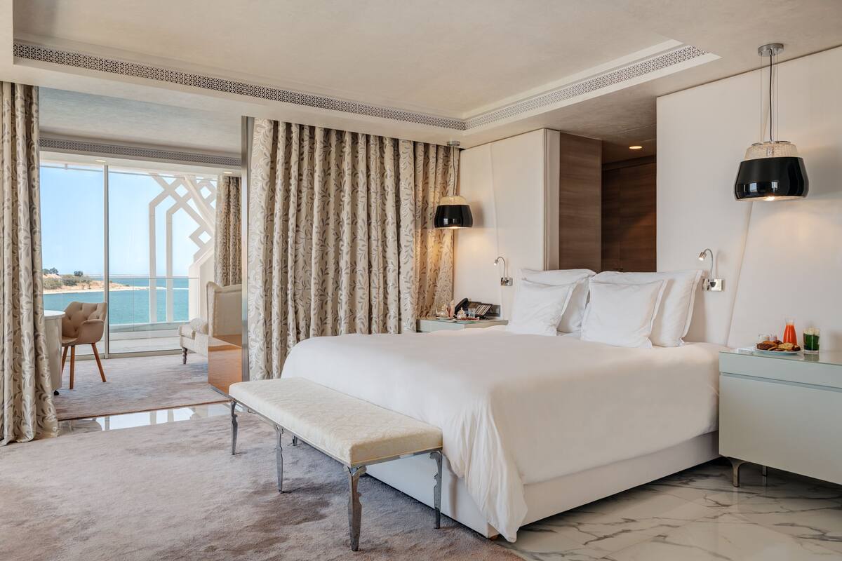A spacious bedroom with a large bed, a bench, a desk, and a view of the sea.
