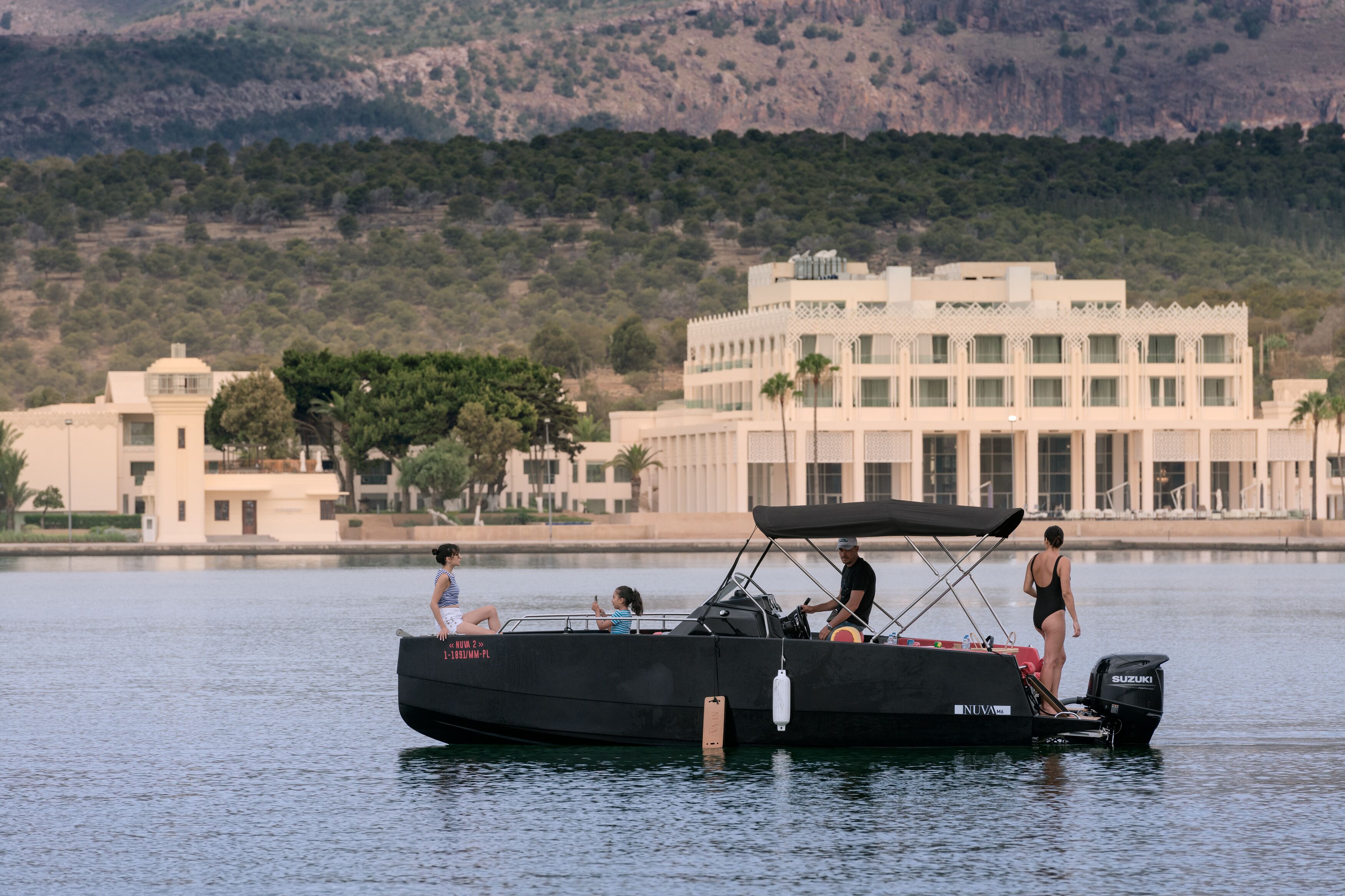 A boat with a black canopy and a Suzuki engine, docked near a building with a white facade and multiple windows.