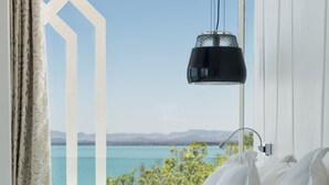 A hotel room with a view of a lake, a bed with white pillows, a bedside table with a blue vase, a white vase with flowers, and a black pendant light. - Marchica Lagoon Resort (Beni Ansar)