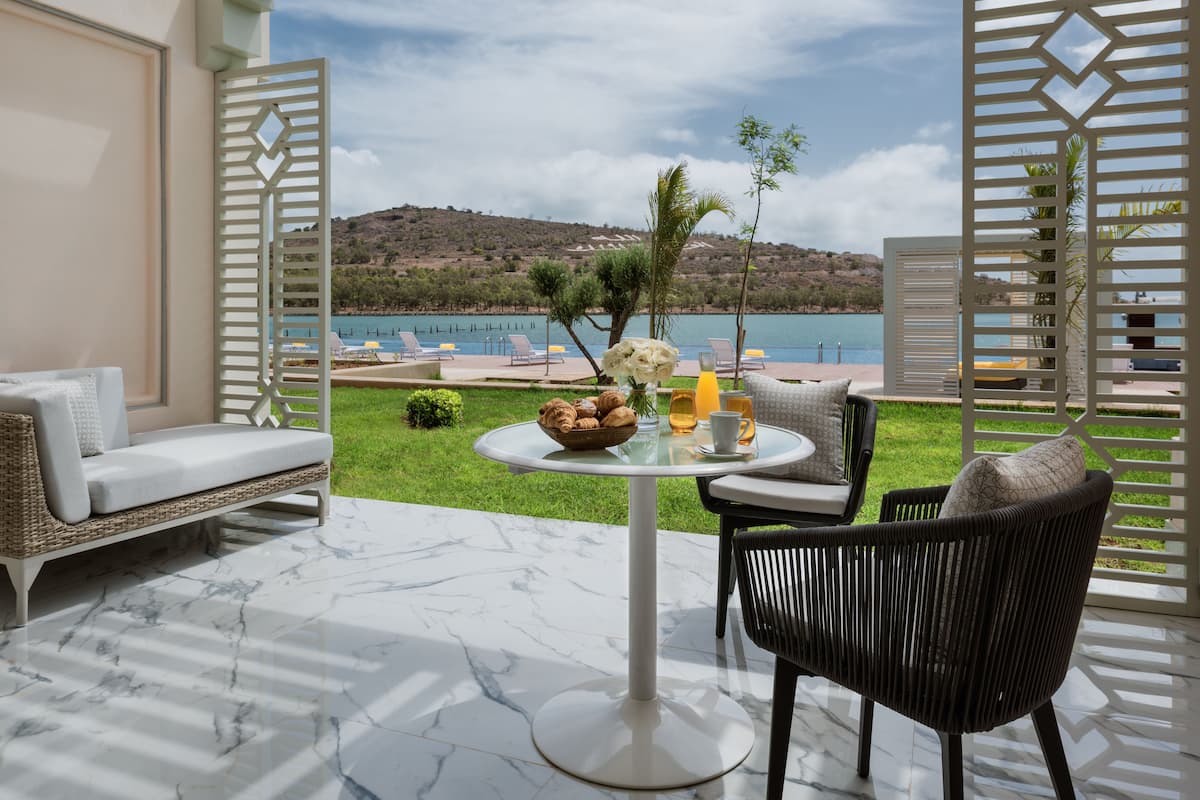 A terrace with a white sofa, a dining table with a bowl of bread and flowers, and a view of a lake and hills.