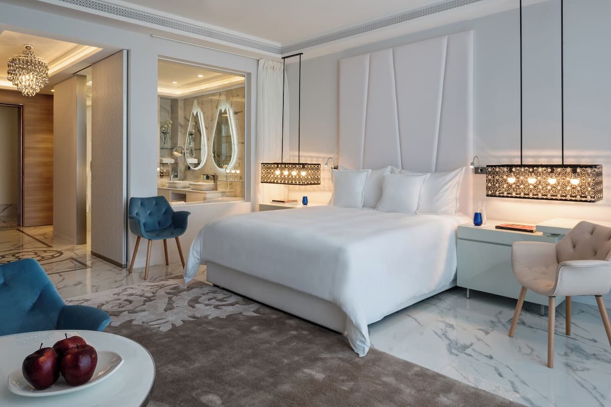 A modern bedroom with a large bed, a nightstand, a vanity with mirrors, and a chandelier.