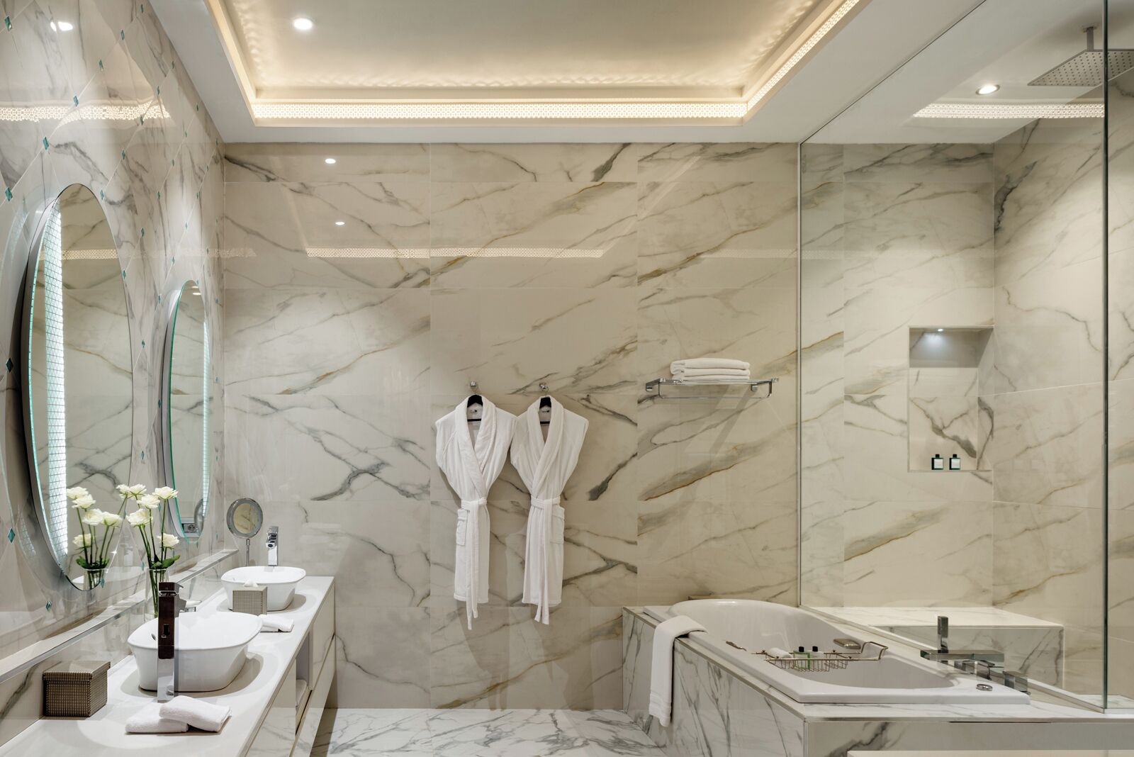Un baño moderno con paredes de mármol, un espejo grande, dos albornoces blancos, una bañera y una zona de ducha con mampara de vidrio.