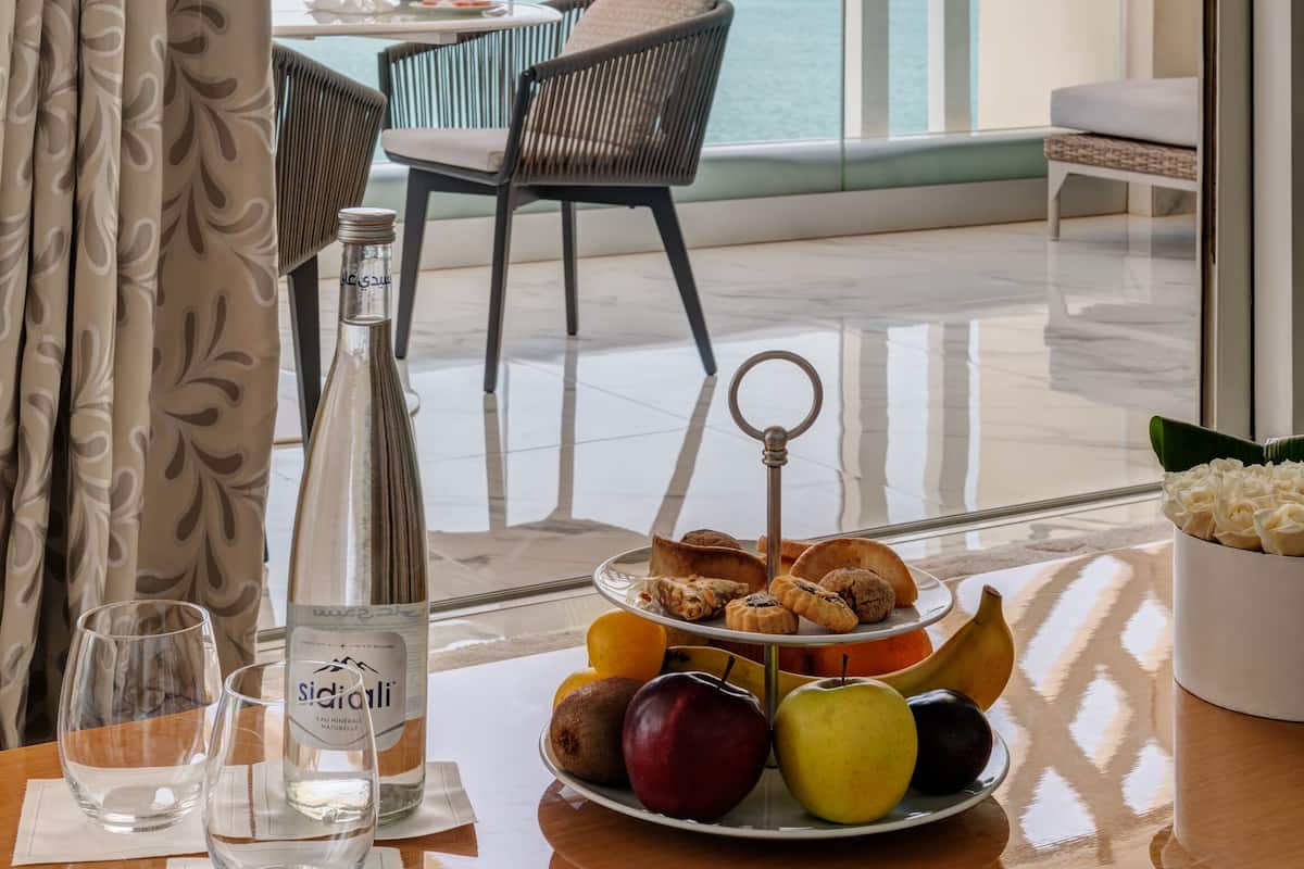 A dining table with a fruit platter, a bottle of water, and cutlery, overlooking a balcony with a view of the sea.