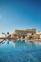 A hotel with a large swimming pool, palm trees, and a clear blue sky.