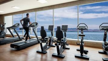 A gym with treadmills, elliptical machines, and stationary bikes, with a large window overlooking a body of water.