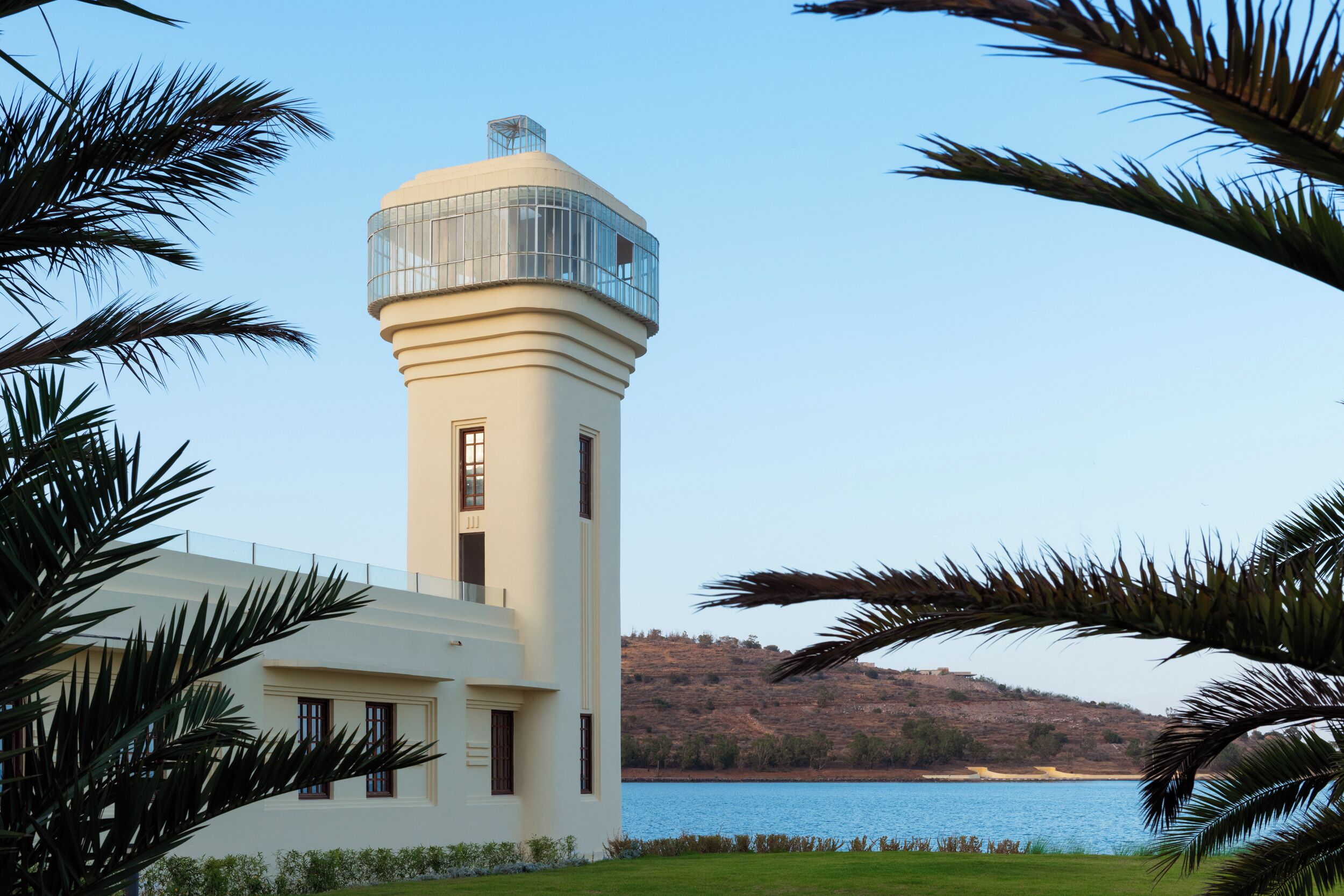 Un faro con una torre cilíndrica y un balcón, rodeado de palmeras y una masa de agua.