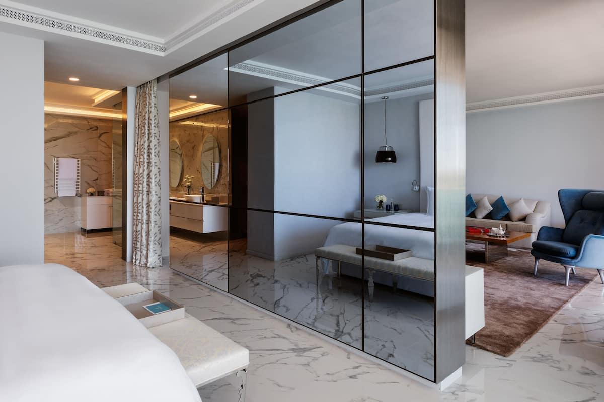 A modern hotel room with a marble floor, a large mirror, a bed, a seating area, and a view of the bathroom.
