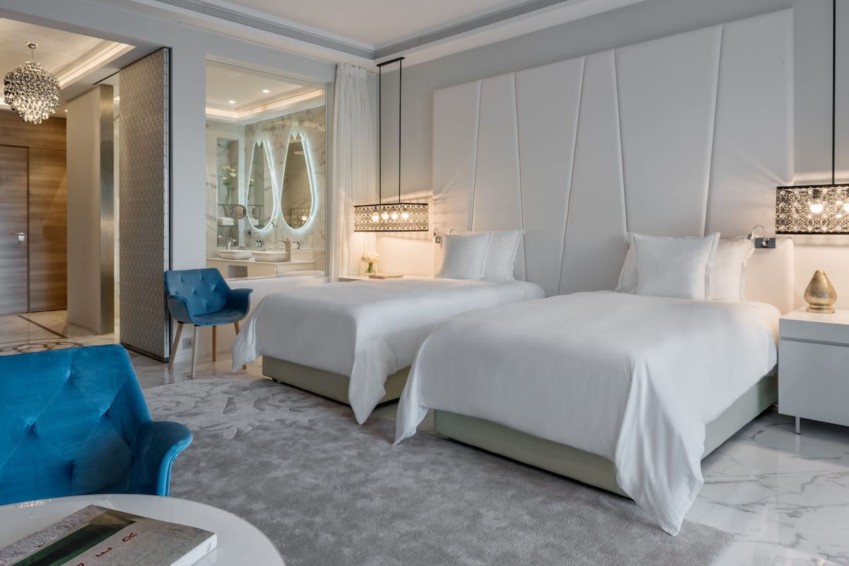 A modern hotel room with two beds, a blue armchair, a round coffee table, and a large mirror.