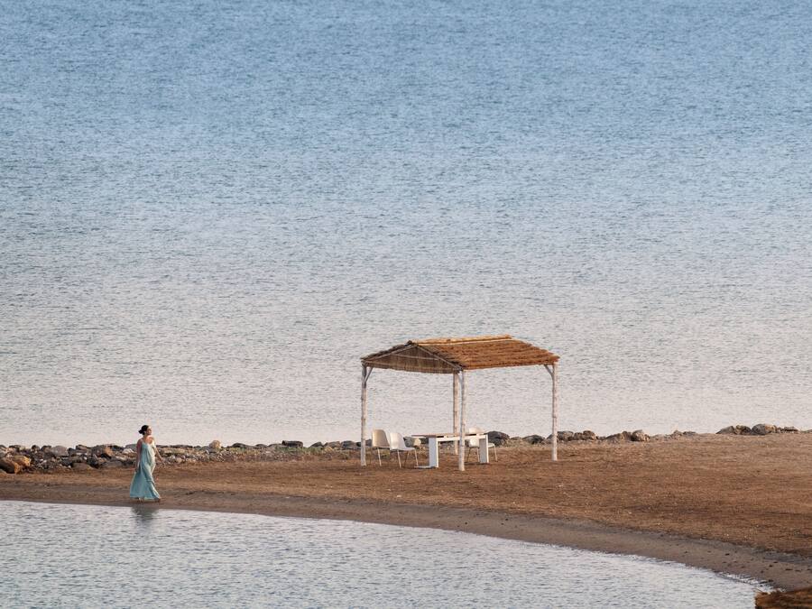 En strand med en træpavillon, en kvinde i en blå kjole, og en stenet kystlinje.