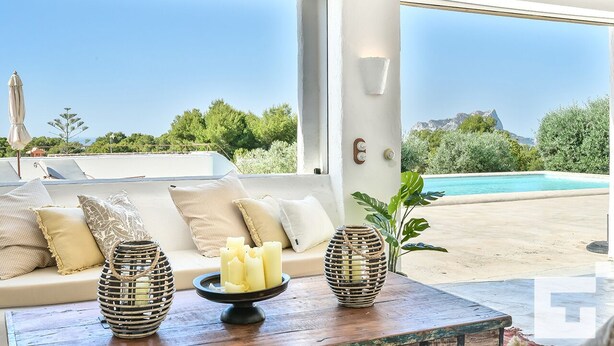 Una habitación iluminada por el sol con un sofá, velas y vistas a una piscina y montañas.