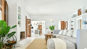 Una sala de estar moderna con un sofá gris, una mesa de centro y una planta grande. Hay una estantería con libros y objetos decorativos. Las paredes son blancas y el piso de madera.