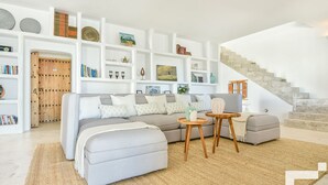 Una sala de estar moderna con un sofá, un reposapiés y una mesa de centro, rodeada de estanterías blancas con varios objetos decorativos.