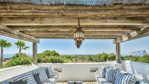 Un área de estar al aire libre con cojines a rayas y un farol colgante, con vistas a un paisaje pintoresco con vegetación y una vista distante del mar.