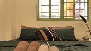 Una cama bien hecha con una colcha verde y varias almohadas, una cesta de mimbre con toallas dobladas y una ventana con marco verde y vistas al exterior.