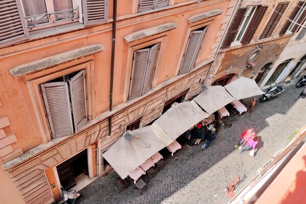 A street-level view of a building with multiple balconies, outdoor seating with umbrellas, and a cobblestone street.