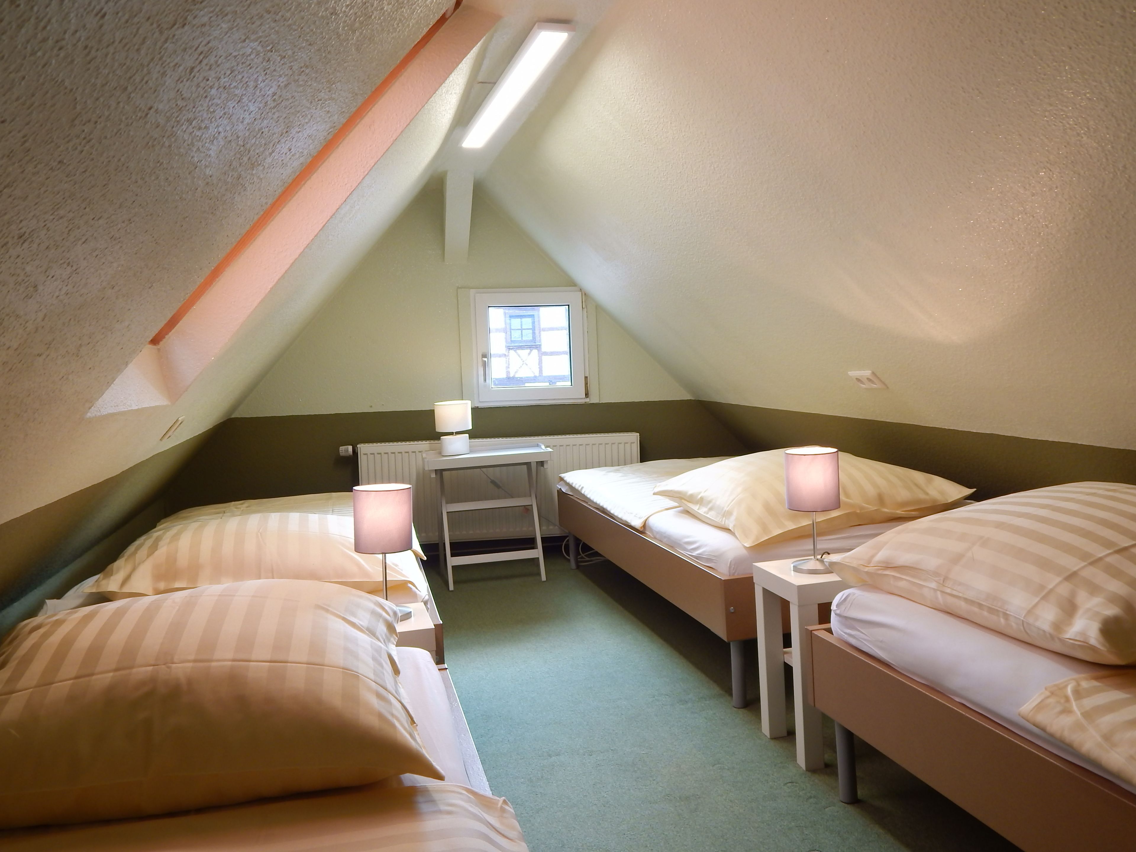Une petite chambre mansardée de forme triangulaire, avec deux lits simples, un bureau et une fenêtre.